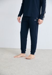 Námornícky modrý top s dlhým rukávom a zodpovedajúce joggerové nohavice z jemnej látky. Top má kontrastné bočné pruhy; joggerové nohavice majú logo na bokoch.