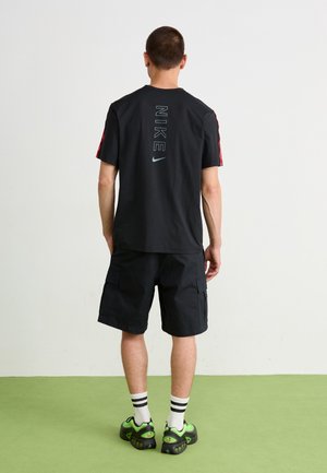 Mężczyzna stojący na zielonej podłodze zwrócony twarzą do białej ściany, ubrany w czarną koszulkę Nike z pionowym logo, czarne krótkie spodnie cargo, białe skarpetki w paski oraz zielone trampki.