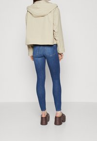Person iført en beige hættejakke med kort længde, blå skinny jeans og brune platform slip-on sko, der står foran en ensfarvet hvid baggrund.