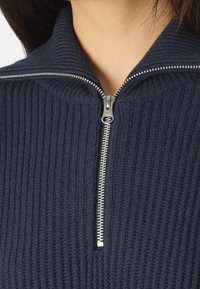 Marineblauwe ribgebreide trui met een hoge kraag en een zilveren ritssluiting aan de voorkant, met een textuurdesign van de stof.