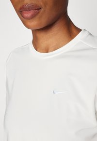 Camiseta deportiva blanca de textura lisa, con un pequeño logo plateado de Nike en el lado izquierdo del pecho y cuello redondo.