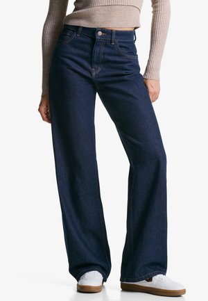 Vrouw draagt donkerblauwe hooggetailleerde wijde jeans, beige geribde top met lange mouwen en witte sneakers met bruine zolen, staand tegen een witte achtergrond.