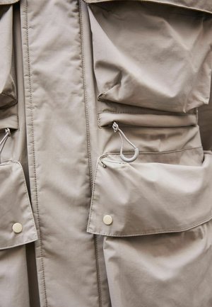 Beige letvægtsjakke med flere lomme-patch, trykknapper og snoretræk detaljer. Glat overflade og vandafvisende materiale.