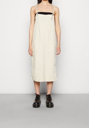 Femme portant une robe midi beige sans manches avec de fines bretelles par-dessus un haut bandeau noir et des bottes noires lacées, debout contre un mur blanc.