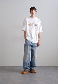 Valge ülipikk t-särk värvilise graafika ja "SHIRT" tekstiga. Seljas helesinised lõtva lõikega teksad ja beežid libisemisega kingad. Minimalistlik taust.