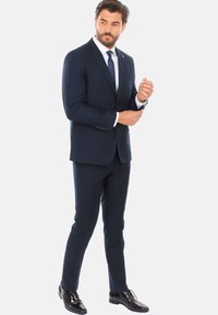 Abito blu navy con vestibilità su misura, giacca monopetto e revers a punta. Camicia bianca e cravatta scura. Scarpa in pelle nera lucida.