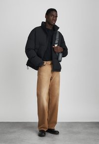 Svart puffjacka med hög krage, kombinerad med en svart skjorta och ljusbruna vida byxor. Modellen håller en gråaxelväska.