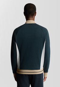 Pullover aus dunkelgrünem Stoff mit beigen Akzenten, ausgestattet mit einem Rundhalsausschnitt, gerippten Bündchen und kontrastierenden weißen Seitenpaneelen. Glatte Textur.