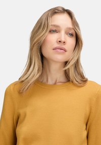 Mustársárga kötött pulóver kerek nyakkal és hosszú ujjakkal. Puha tapintású, egyszerű dizájnnal, bordázott aljjal és mandzsettákkal.