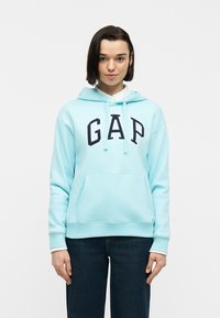 Νεαρή γυναίκα με γαλάζιο φούτερ GAP και σκούρο τζιν, στέκεται με τα χέρια χαλαρά στο πλάι μπροστά σε μονόχρωμο φόντο.