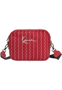 Červená látková crossbody taška s vlnitými bílými pruhy a podpisovým detailem. Obsahuje nastavitelné ramenní popruhy a zipové uzavření.