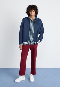 Navy bomber jacket with white striped cuffs over a green and blue striped shirt, paired with maroon sweatpants and white sneakers.