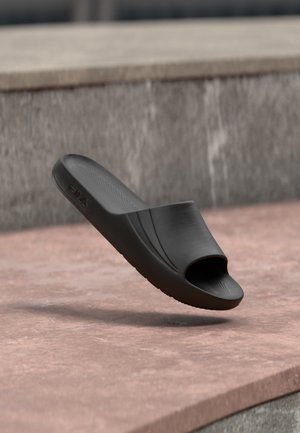 Sandalo nero tipo ciabatta con plantare testurizzato e punta aperta, sospeso sopra una superficie di pietra con sfondo di muro di pietra grigia.