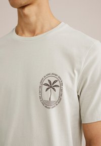 Lysegrå bomuld t-shirt med rund halsudskæring, prydet med et brunt palmetræ-grafik og tekst, der omkranser det. Glat tekstur.