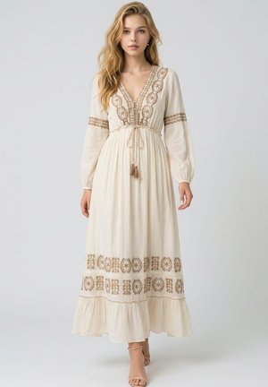 Joven mujer que lleva un vestido largo color crema con patrones bordados en marrón y mangas largas, de pie frente a un fondo claro y sencillo.