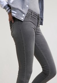 Osoba oblečená v šedých skinny džínách a šedé varsity bundě, ruka odpočívá v zadní kapse, na čistém světlém pozadí.