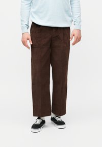 Ανδρικό παντελόνι καφέ από κορδόνι με φαρδύ πόδι, σε συνδυασμό με μαύρα αθλητικά παπούτσια, λευκές κάλτσες και ανοιχτό μπλε πουλόβερ σε λευκό φόντο.