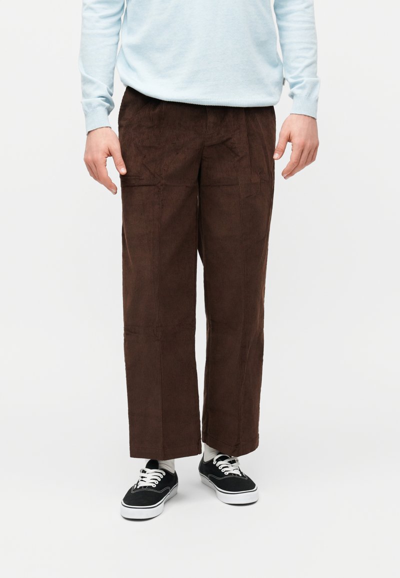 Ανδρικό παντελόνι καφέ από κορδόνι με φαρδύ πόδι, σε συνδυασμό με μαύρα αθλητικά παπούτσια, λευκές κάλτσες και ανοιχτό μπλε πουλόβερ σε λευκό φόντο.