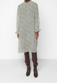 Vestido abotonado en patrón de pata de gallo en blanco y negro, con mangas largas y puños con volantes, y una cintura entallada, combinado con botas marrones hasta la rodilla.
