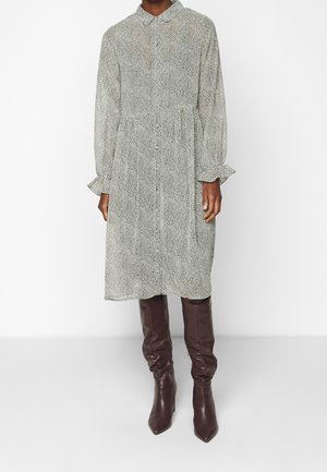 Button-up dress in black and white houndstooth pattern, featuring long sleeves with ruffled cuffs and a cinched waist, paired with brown knee-high boots.