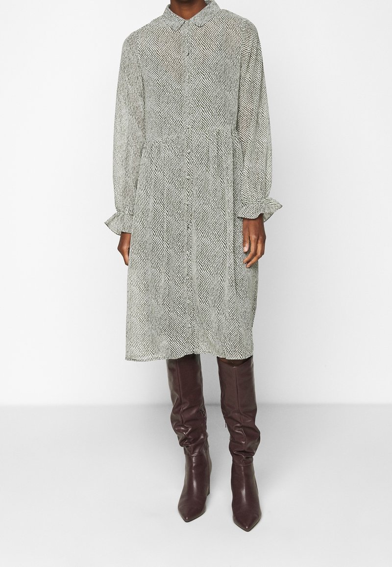 Vestido abotonado en patrón de pata de gallo en blanco y negro, con mangas largas y puños con volantes, y una cintura entallada, combinado con botas marrones hasta la rodilla.