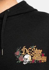Sudadera negra con un gráfico de calavera y rosa en rojo y verde. Presenta "DC Shoe Company" en texto amarillo negrita; tela suave y texturizada.