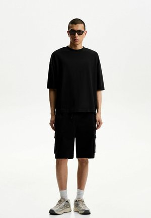 Zwarte katoenen t-shirt gecombineerd met zwarte cargo shorts. Het model draagt een zonnebril en beige sportschoenen met grijze accenten en witte sokken.