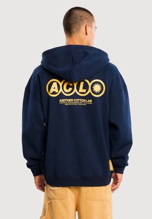 Mann trägt dunkelblaue Kapuzenjacke mit gelbem „ACLO Another Cotton Lab“-Schriftzug und Symbolen auf dem Rücken, kombiniert mit beigen Hosen, von hinten gesehen.