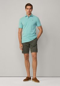 Svetlomodrá polo košeľa s krátkym rukávom v kombinácii s olivovo zelenými šortkami. Model má na sebe hnedé semišové slip-on topánky. Minimalistický dizajn.
