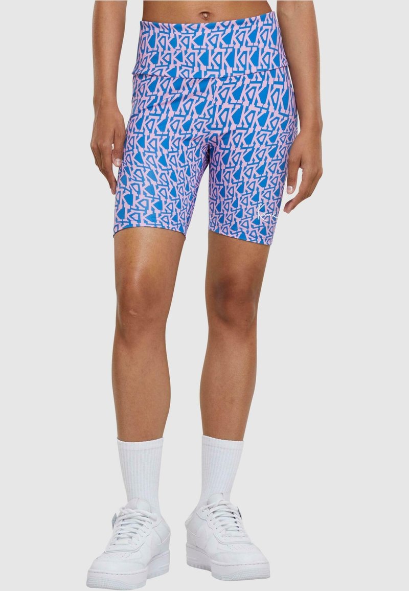 Personne portant un short de cycliste bleu et rose à motifs avec des lettres "KK" répétées, des chaussettes blanches et des baskets blanches sur un fond uni.