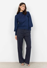 Maglione blu navy con collo alto e maniche a palloncino, abbinato a pantaloni scuri a righe. Le scarpe del modello hanno una finitura metallica.