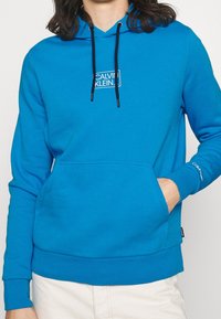 Sudadera turquesa con bolsillo frontal, cordones negros y logo blanco de Calvin Klein. Puños y dobladillo acanalados, fabricada con tejido suave.