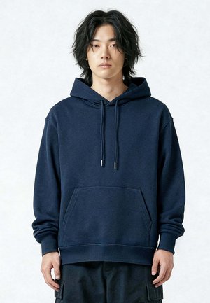 Jonge persoon met halflang zwart haar, gekleed in een loszittende donkerblauwe hoodie en zwarte cargobroek, staand tegen een effen achtergrond.