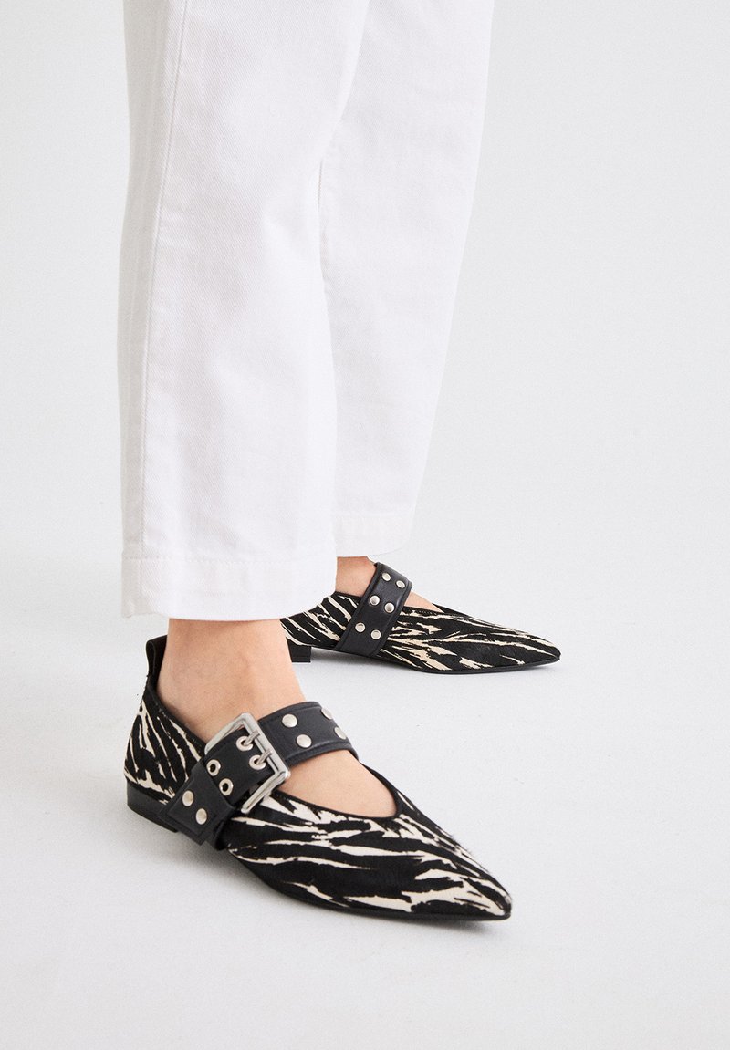 Bailarinas de punta en tela de estampado cebra en blanco y negro, con una ancha tira negra con tachuelas metálicas, usadas con pantalones cropped blancos.