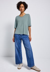 T-shirt vert clair à col en V avec des manches courtes, associé à un jean bleu à jambes larges et des baskets beiges. Texture lisse, coupe décontractée.