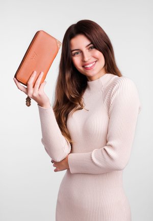Brown leather wallet with a textured corner, held by a model in a light pink ribbed sweater, displayed against a neutral background.