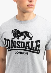 Pilkas medvilnės marškinėliai su juodu liūto grafikų ir „LONSDALE LONDON“ tekstu. Trumpomis rankovėmis, apvaliu iškirpimu, lygia tekstūra.