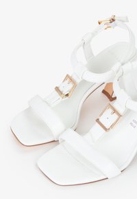 Hvide læder højhælede sandaler med firkantede tåspidser, udstyret med justerbare stropper og spænder i guldfarve; glat tekstur med minimalistisk design.