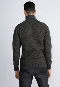 Maglione grigio scuro con colletto alto, orlo a costine e motivo testurizzato, abbinato a jeans neri, fotografato di schiena su uno sfondo chiaro.