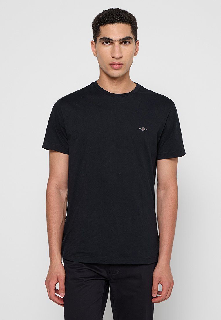 Młody mężczyzna z krótkimi, kręconymi włosami, ubrany w prostą, czarną koszulę z krótkim rękawem i czarne spodnie, stojący na białym tle.