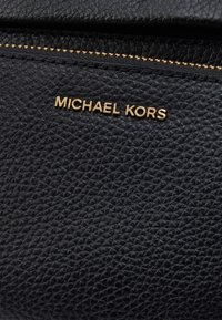 Melnā teksturētā ādas materiāls ar zelta "MICHAEL KORS" zīmolu. Tam ir rāvējslēdzēja aizsardzība un izteiksmīga ģeometriska raksts uz virsmas.