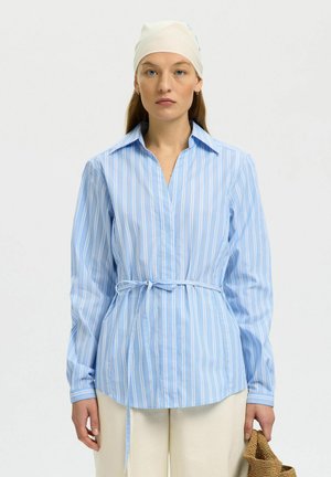 Žena v svetlomodrej prúžkovanej blúzke s opaskom v páse, bielou šatkou na hlave, krémovými nohavicami a drží v pravej ruke pletenú tašku.