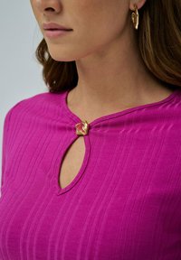 Top de color fucsia con ribetes y escote en forma de llave, que presenta un exclusivo detalle de botón dorado en el centro. Textura suave y diseño ajustado.