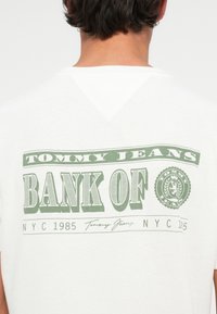 Személy fehér pólót visel, melynek hátulján a "Tommy Jeans Bank of NYC 1985" felirat és egy zöld, kör alakú embléma található.