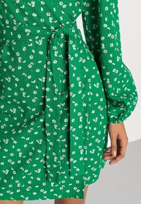 Vestido floral verde con una tela texturizada, mangas largas y un detalle de cintura atada, con pequeños patrones de flores blancas y marrones en todo el diseño.