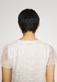 Persona con capelli neri corti, che indossa piccoli orecchini a cerchio e una maglietta trasparente beige con un motivo, con il logo rosa "Desigual" sulla parte superiore della schiena.