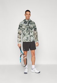 Lettere, mønstrede jakke i grøn og beige, parret med sorte shorts. Holder en tennisracket og bærer hvide sneakers.