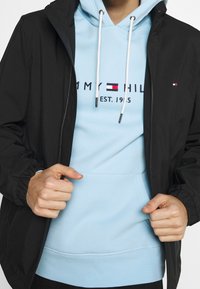 Černá lehká bunda přes světle modrou mikinu s kapucí. Mikina má logo a bílé šňůrky na stažení. Bunda má elastické návleky a jednoduchý design.