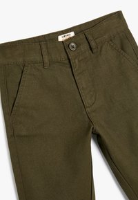 Olijfgroene katoenen kinderbroek met een knoopsluiting, diagonale voorkruiszakken en een gladde textuur. Label zichtbaar bij de tailleband.