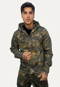 Giacca mimetica in tonalità verdi e marroni, con due tasche frontali, chiusura a bottoni, bordi sfrangiati e cappuccio.